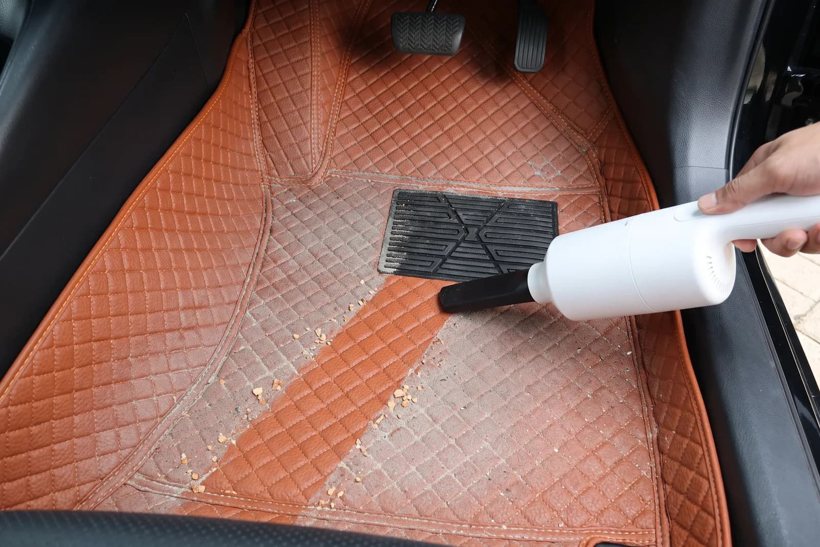 A handheld vacuum leaving a clean trail through crumbs and dust on the driver's Porsche Orange 3D car mat
