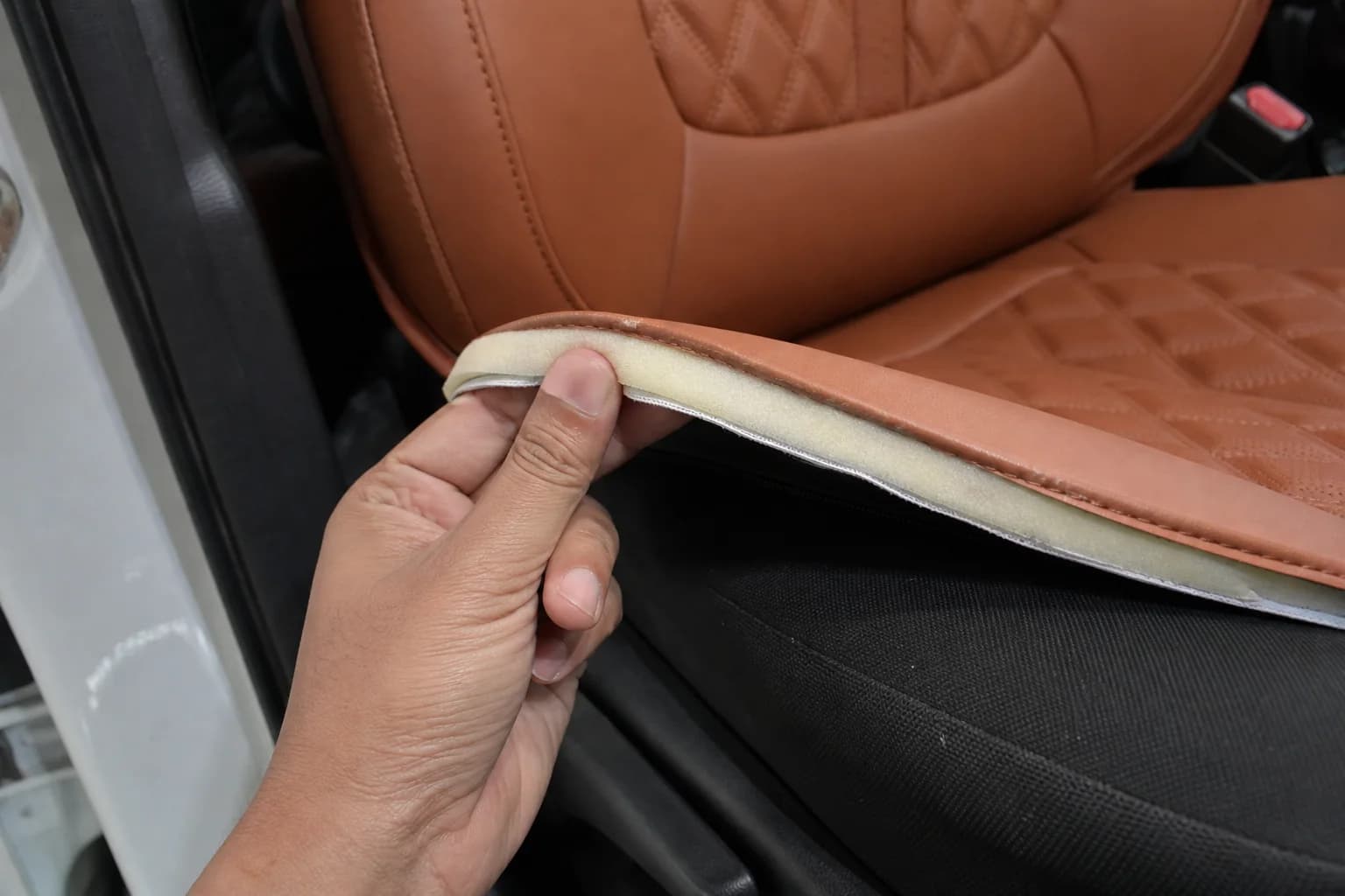 A hand lifting the corner of a tan quilted seat cover to show the thin foam layer bonded under the leatherette